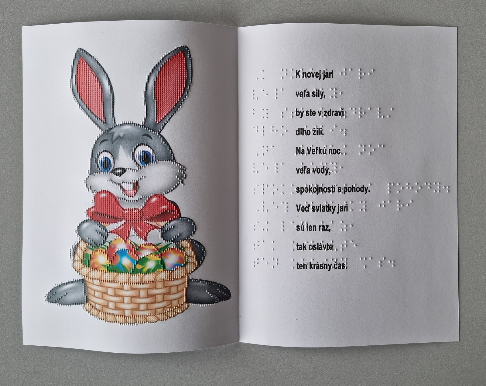 Fotografia pohľadnice, na ľavej strane je farebný obrázok v bodovej grafike (zajko s červenou mašľou a košíčkom s vajíčkami), na pravej strane text v braili a čiernotlači.