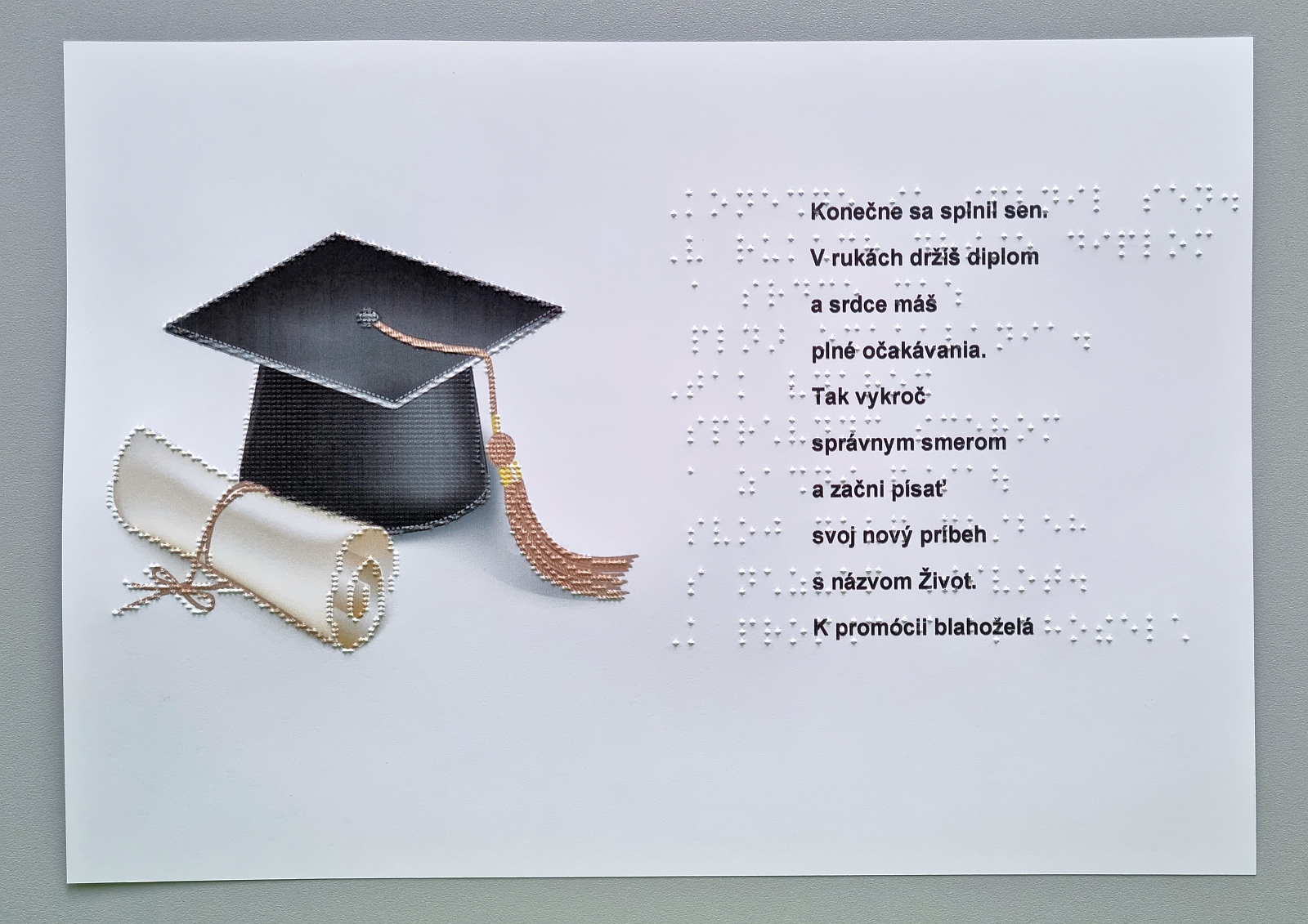 Fotografia pohľadnice k promócii, na ľavej strane je promočná čiapka s visiacimi strapcami a tubusom s diplomom, na pravej strane blahoželanie v braili.