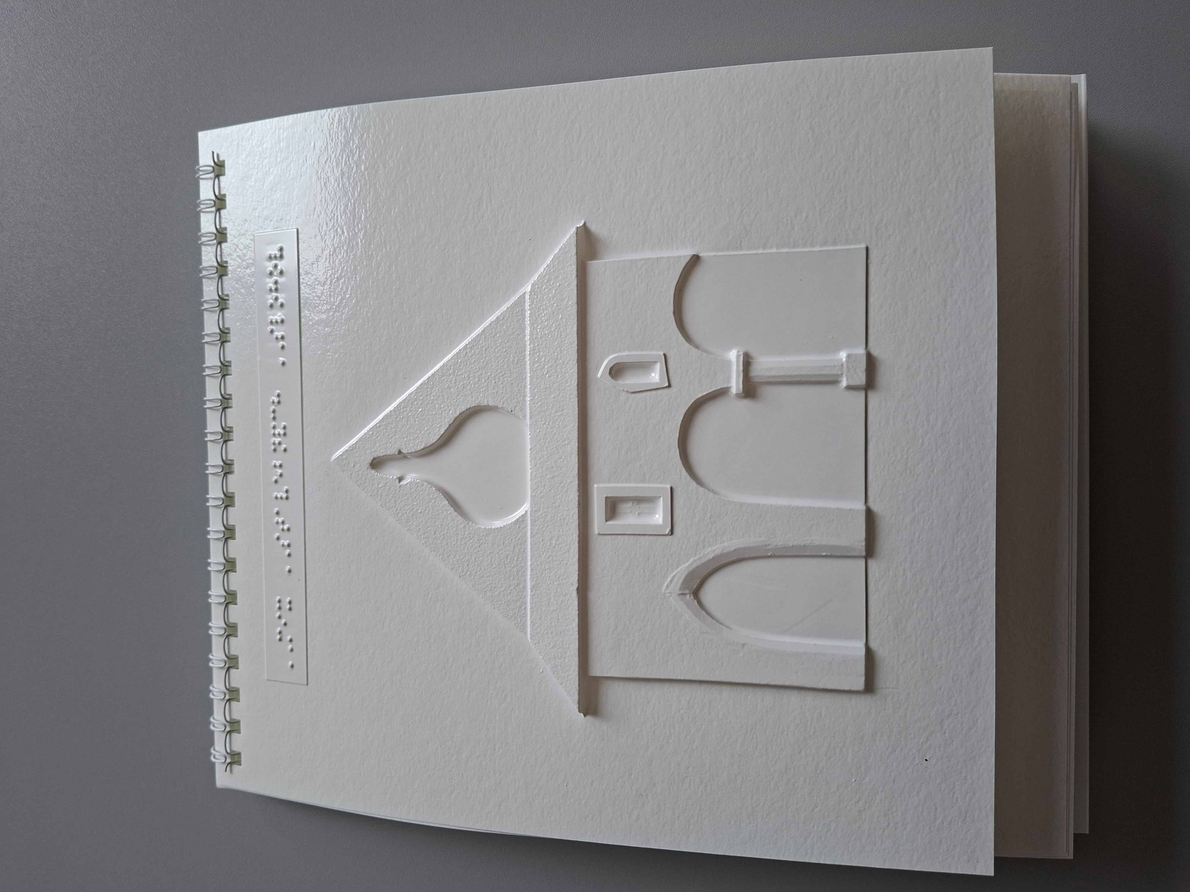 Biely list v špirálovej väzbe s reliéfnym obrázkom budovy pripomínajúcej kostol alebo kaplnku: trojuholníková strecha, hore malý tvar ako vežička alebo zvon, na fasáde oblúkový vstup a ďalšie oblúky. Nad obrázkom je krátky riadok braillovho písma.