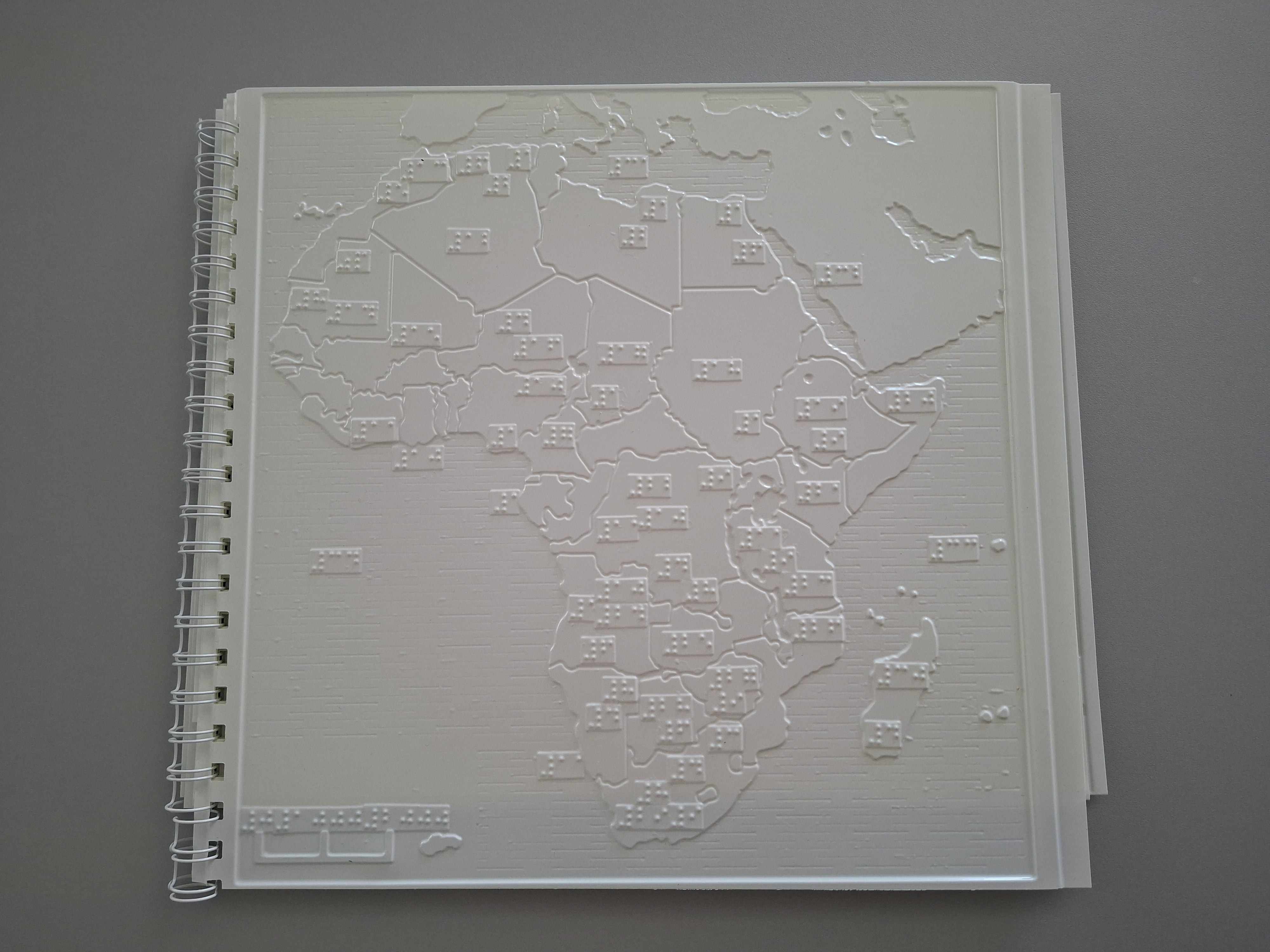 Reliéfna hmatová mapa Afriky v plastovom viazanom zošite leží otvorená na šedej podložke. Na stránke sú vyvýšené obrysy afrického kontinentu rozdelené na jednotlivé štáty tenkými čiarami. Pri každom štáte sú malé obdĺžnikové políčka s bodkami v Braillovom